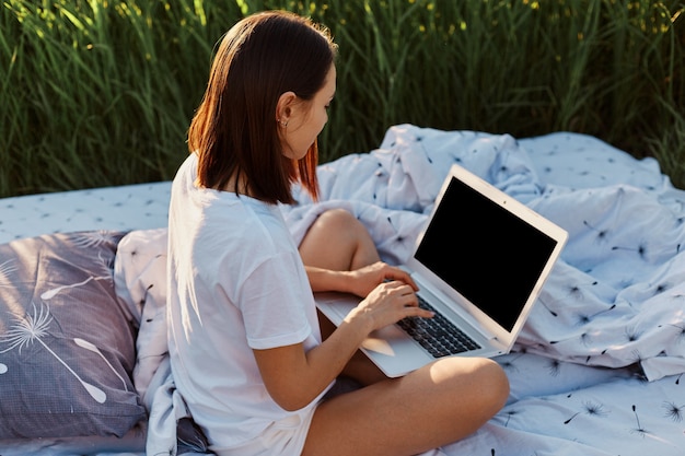 Vista laterale della donna dai capelli scuri che indossa una maglietta casual bianca seduta con le gambe incrociate e tiene in mano un lap top con schermo vuoto, libero professionista che lavora online su un letto morbido nel prato.