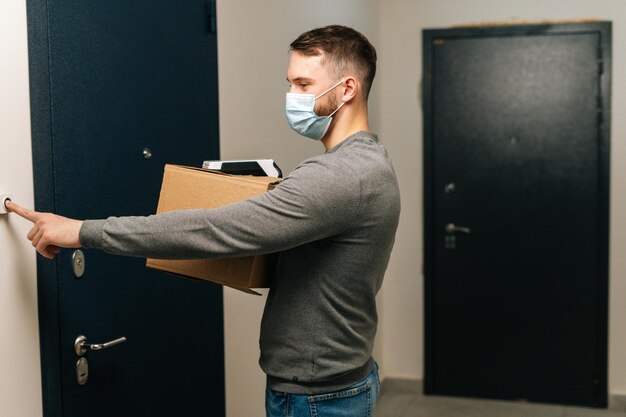 Side view of delivery man wearing medical mask holding cardboard box and contactless payment POS wireless terminal for card paying ringing doorbell
