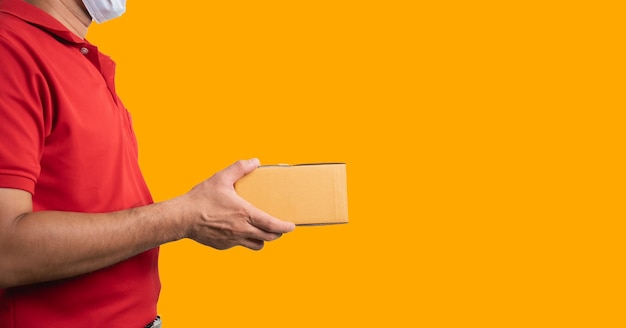 Foto vista laterale operaio di consegna che indossa maschera chirurgica e guanti medici in uniforme rossa isolato su sfondo giallo tenere cassette dei pacchi per l'invio di pacchi per posta carta da parati larga