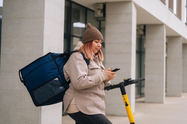 Side view of delivery woman with thermo backpack using navigation app on mobile phone riding order on electric scooter