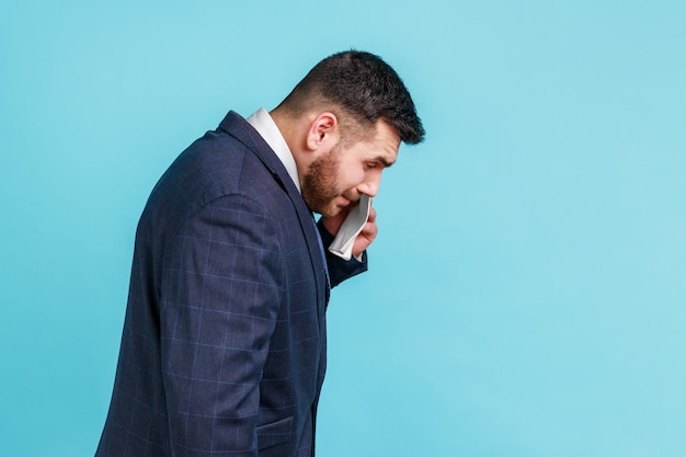 Side view of depressed brunette man with beard wearing official style suit wipe tears with a handkerchief, looking desperate, crying. Indoor studio shot isolated on blue background.