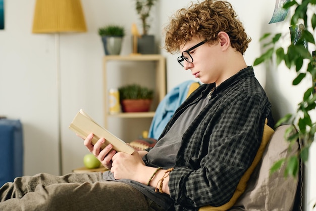 안경 과 캐주얼 의류 를 입은 부지런 한 십대 소년 의 측면 시선 책 읽기