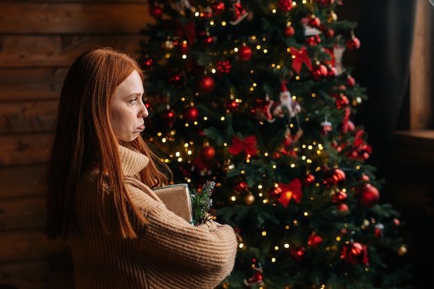 Side view of dissatisfied young woman holding Christmas gift box on background of xmas tree