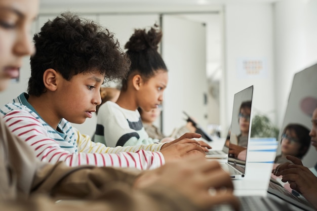 Photo side view at diverse group of children sitting in row at school classroom and using computers