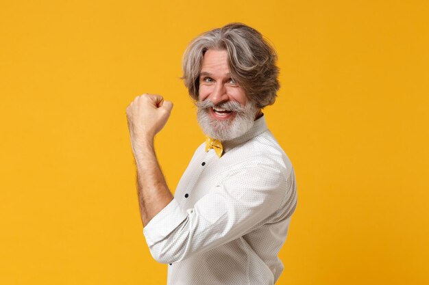 Side view of elderly gray-haired mustache bearded man in white shirt bow tie posing isolated on yellow orange background in studio. people lifestyle concept. mock up copy space. doing winner gesture