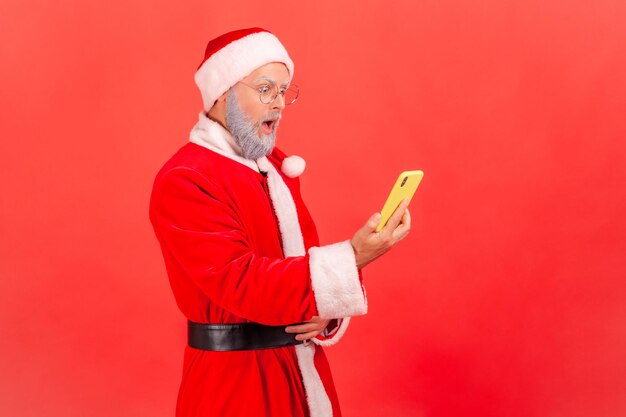 Foto vista laterale di un uomo anziano con la barba grigia che indossa il costume di babbo natale in piedi con la bocca aperta e