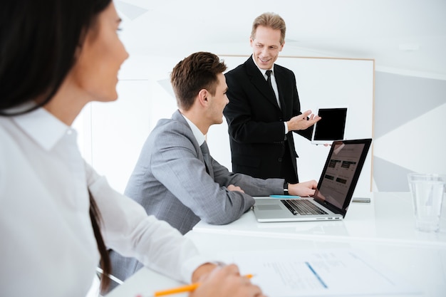 Side view of Elderly smiling business man showing blank tablet computer screen to his colleagues on conference