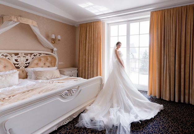 Side view of elegant bride stands at the window of a hotel room and looks into the camera