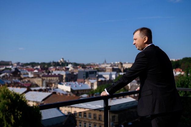 Side view of elegant bridegroom wearing in luxury black suit standing on terrace leaning on transparent railing looking and enjoying of amazing view on ancient downtown during wedding in summer day