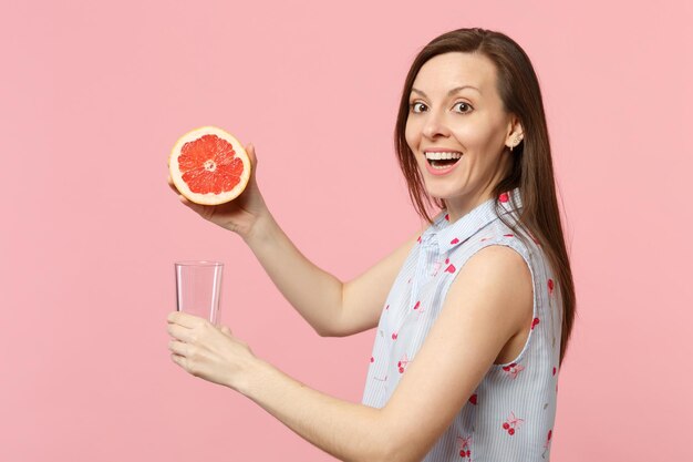 Side view of excited young girl in summer clothes holding half of fresh ripe grapefruit glass cup isolated on pink pastel background. people vivid lifestyle relax vacation concept. mock up copy space