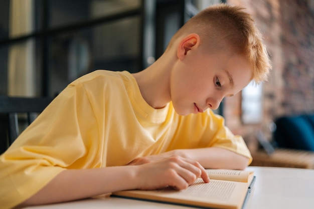 Side view face of adorable pupil schoolboy reading paper book following lines with finger along page sitting at desk in living room