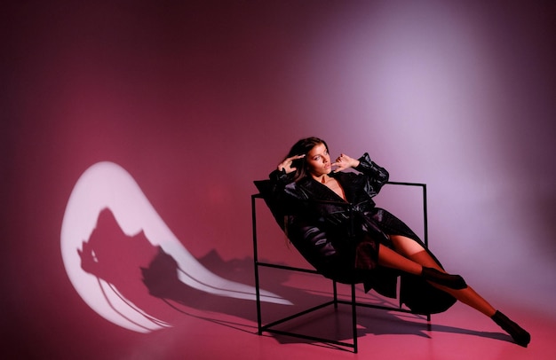 Side view of fashionable female model wearing black leather\
trench coat and heels posing and photographing for magazine cover\
while sitting on metal chair in studio with red spotlight\
illumination