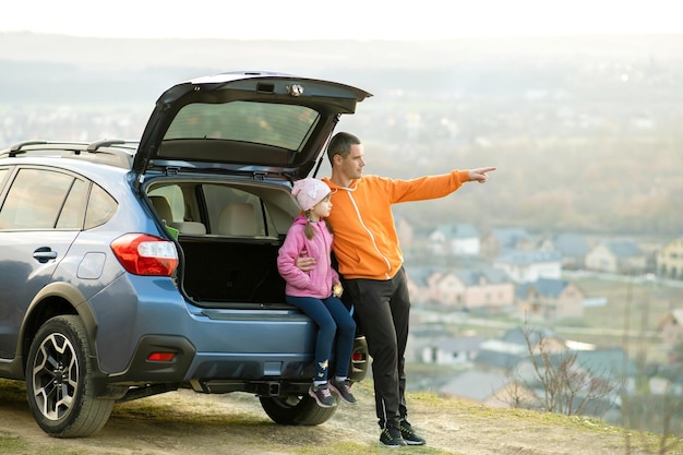 Вид сбоку отца с маленькой дочерью, стоящей возле автомобиля с открытым багажником и наслаждающейся природой. Концепция выходных с семьей.