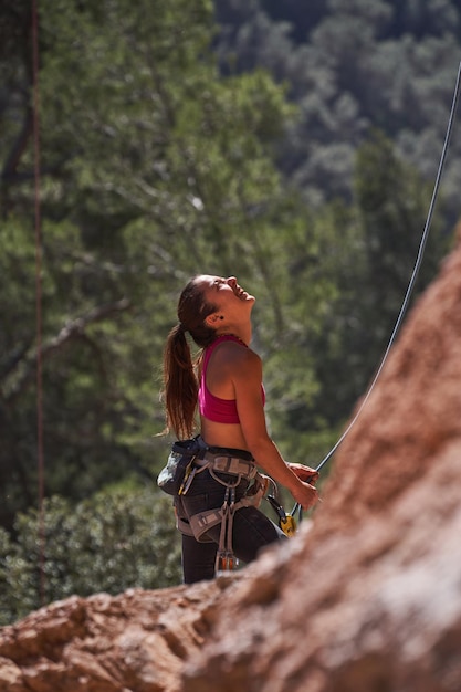 Side view of female climber in safety harness looking up while standing near rocky cliff against green trees in nature