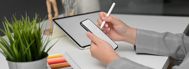 Side view of female designer working with mock-up tablet while looking for information on smartphone