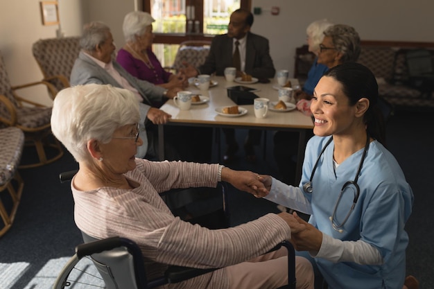 Side view of female doctor holding hands and talking with disable senior woman at nursing home