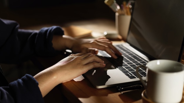 Side view of female hand typing on laptop on wooden table with coffee cup