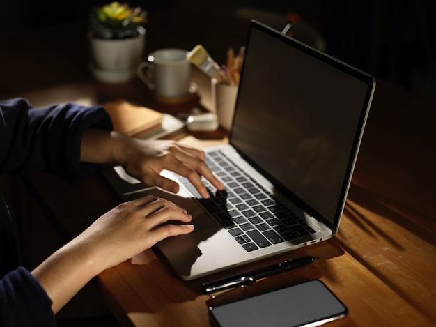 Вид сбоку женской руки, печатающей на ноутбуке на деревянном столе со смартфоном и принадлежностями