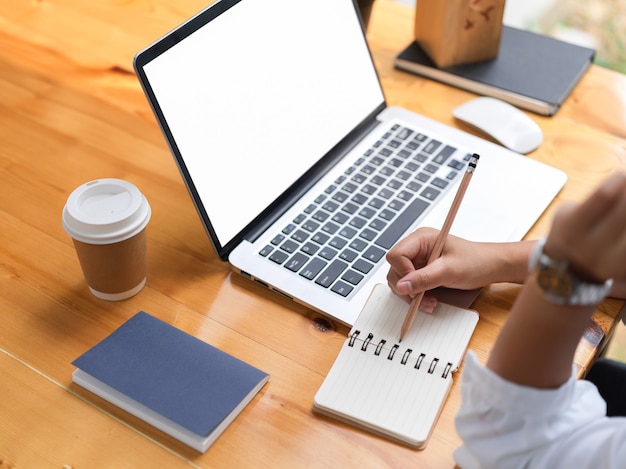 Vista laterale della mano femminile che scrive sul taccuino in bianco mentre lavora con il computer portatile sul percorso di residuo della potatura meccanica della tavola in legno