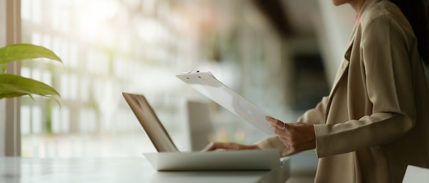 Photo side view of female office worker working with laptop and paperwork on table in glass wall office