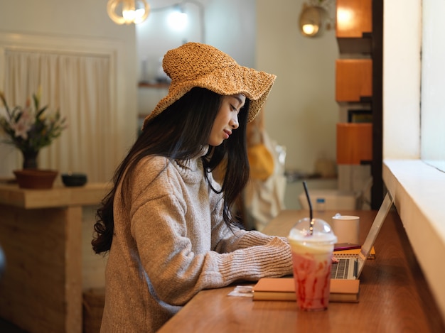 Vista laterale della donna in maglione e cappello lavorando con laptop e articoli di cancelleria mentre è seduto al bancone della caffetteria