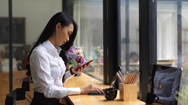 Side view of female using smartphone while working with digital tablet on wooden bar in cafeteria