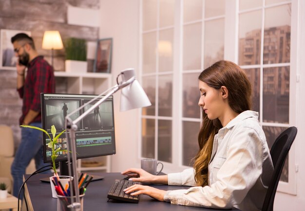 side view of female videographer working on post production of a movie from home . boyfriend in the background.