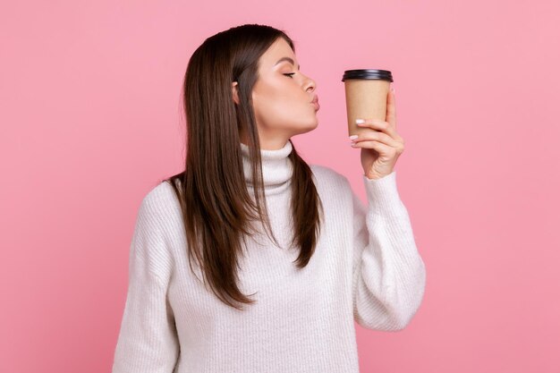 Side view of female with dark hair kissing disposable cup of take away coffee, enjoying beverage, wearing white casual style sweater. Indoor studio shot isolated on pink background.