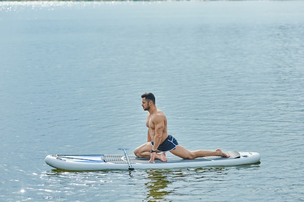 Side view of fit man doing stretching exercises sitting on sup board.