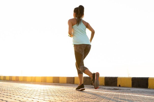 Side view of fitness woman running on a road by the sea near the sea port. Sportswoman training on seaside promenade at sunset.