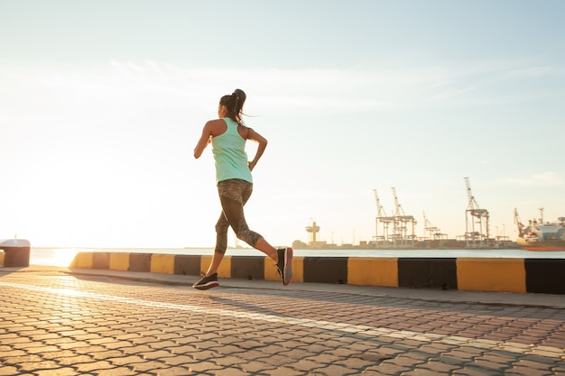 Side view of fitness woman running on a road by the sea near the sea port. Sportswoman training on seaside promenade at sunset.