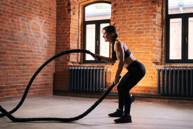 Side view of fitness young athletic woman with strong beautiful body in black sportswear exercising with battle ropes having workout training. Muscular Caucasian female workout out in dark gym.