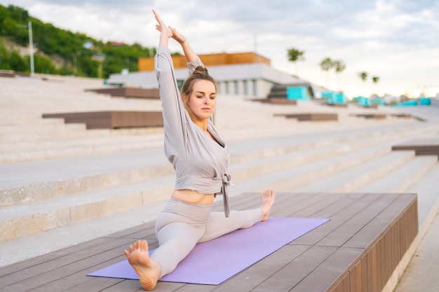 Side view of flexible slim sporty woman wearing sportswear sitting on twine with arms up on yoga mat outdoors