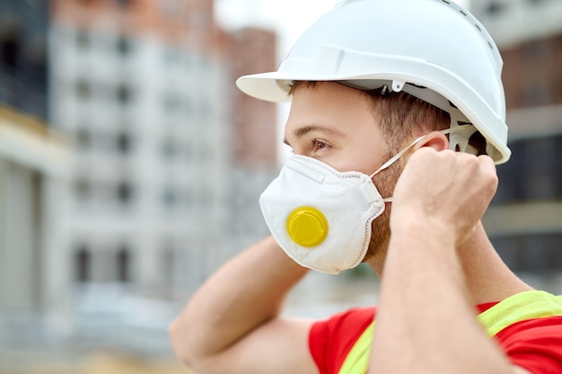 Side view of a focused dark-haired bearded builder in headwear putting on a protective mask outside