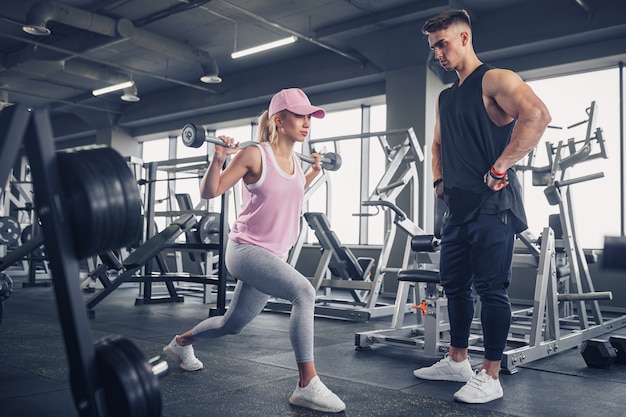 Side view of focused and motivated sporty young blonde girl in sportswear doing legs exercises while handsome muscular personal trainer monitoring her in the gym