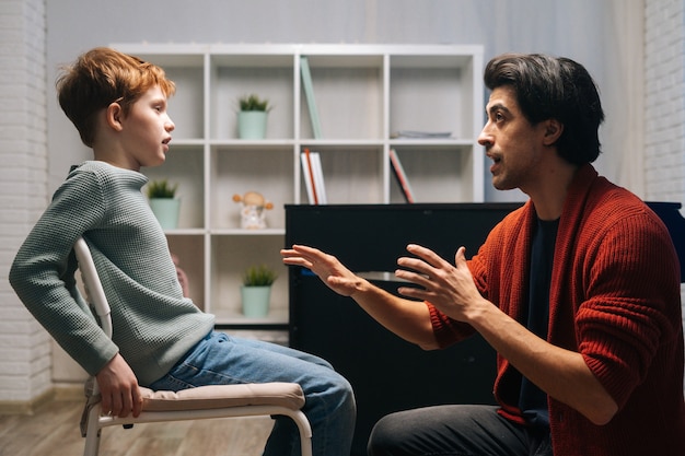 Side view of focused schoolboy sitting on chair listening young music teacher talking and gesturing