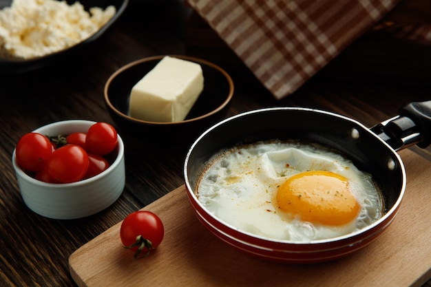 Side view of fried egg in frying pan on cutting board with tomatoes butter cottage cheese on wooden background