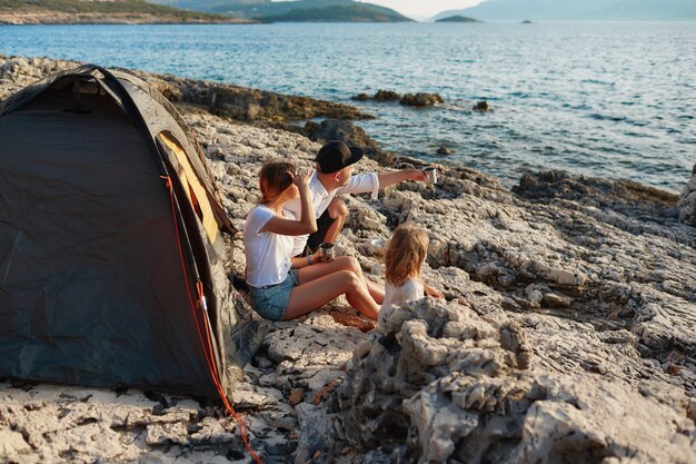 Foto vista laterale della famiglia amichevole che si siede vicino alla tenda alla spiaggia della roccia, mare ammiratore.