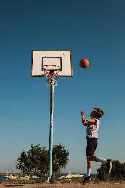 Вид сбоку всего тела мальчика в спортивной одежде, играющего в стритбол, прыгающего, бросая мяч на спортивную площадку