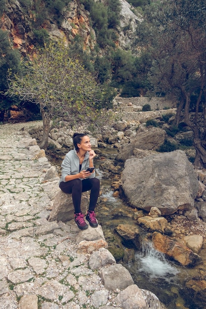 Side view full body of content relaxed woman enjoying trip near streaming creek in mountainous terrain while sitting on rock with smartphone and looking away