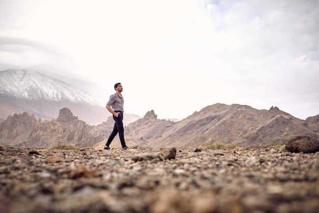 Side view full body ground level of bearded Hispanic male traveler walking with hands in pockets on rocky terrain near mountain Teide and looking away in Tenerife Canary Islands Spain