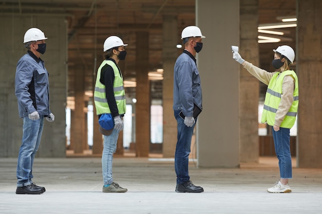 Side view full length of female supervisor measuring temperature of workers with contactless thermometer at construction site, corona virus safety measures