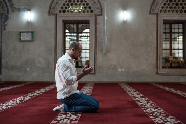 Photo side view full length of man praying while kneeling on carpet in mosque