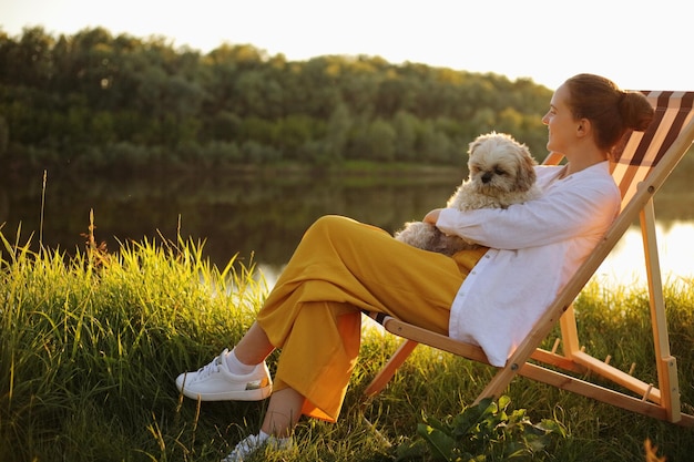 Ritratto a figura intera vista laterale di felice giovane donna adulta che indossa una camicia bianca e il suo cane pechinese seduto al tramonto vicino al fiume godendo di una splendida vista