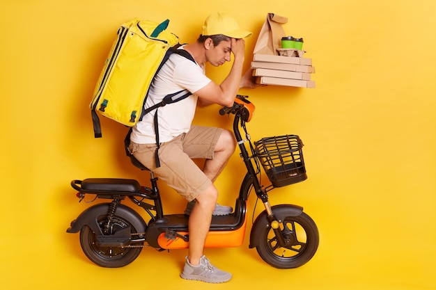 Side view full length portrait of man courier with thermo backpack in white Tshirt and cap delivering pizza and coffee looks tired waiting client long time isolated over yellow background