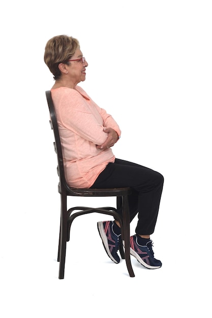 Side view of a full portrait of senior woman sitting on chair dressed in sportswear and smiling on white background