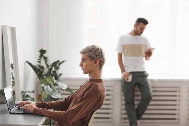 Side view of gay couple everyday life focus on blonde young man\
using laptop while working from home in foreground, copy space