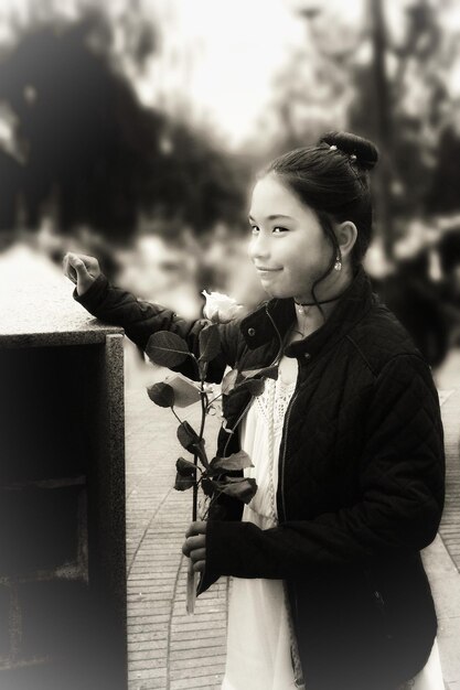 Photo side view of girl holding rose looking away while standing on footpath