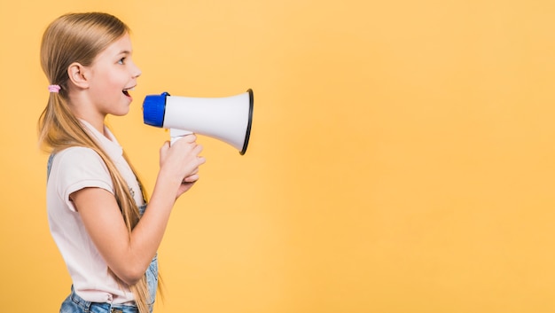 Side view of a girl loudly speaking through megaphone against yellow backdrop