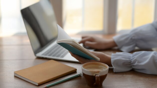 Side view of a girl typing on laptop while looking on her notebook on wooden desk